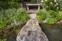Boulder over a pond leading to a stone bench with drystone wall surround, marginal planting of Phalaris arundinacea and Rhododendron