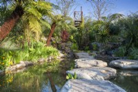 Pond with large stone boulders as stepping stones leading to a rusty spiral staircase viewing platform, marginal planting and Dicksonia antarctica tree ferns 