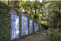 Antique tiled panels adorn an ivy covered wall with trees overhead and a cobbled path in front. Monte Palace Gardens, Madeira. August. Summer
