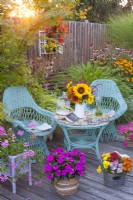 Raised bed planted with Pelargoniums, a wicker container with Impatiens, a bouquet of summer flowers including sunflowers and wicker furniture on decked patio.