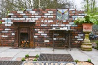 Decorative colorful brick wall with a fireplace, old sewing machine table and  vase with ferns. Fragment of  mixed paving with salvage materials including slabs, bricks, concrete block pavers and pebbles with reclaimed drain covers with decorative  rusted drainage grates and metal grill.  April
Designer: Pam Creed