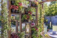 A wooden partition wall with vertical garden in pots planted with Erigeron karvinskianus, Polypodium vulgaris,Heuchera 'Peach Flambe', Trachelospermum jasminoides and decorated with lanterns shelters the outdoor dining area. June