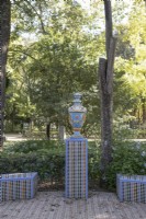 A colourful, glazed urn, on top of a plinth, with curving low, tiled benches decorated in the Mudejar, Islamic style. various shrubs and trees are in the background.  Parque de Maria Luisa, Seville, Spain. September
