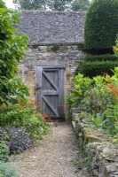 A gravel path leads past a Cotswold stone retaining wall, yew topiary and summer flowers towards a wooden door in a brick wall at The Manor, Little Compton.