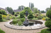 A circular pond with fountain is surrounded by a gravel path and borders with topiary and perennials in The Flower Garden at The Manor, Little Compton.