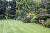 A wooden seat is set into a mixed shrub border alongside a mown lawn at The Manor, Little Compton.