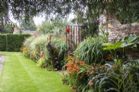 Borders filled with tropical style planting run either side of a wrought iron gate set into an old brick wall at The Manor, Little Compton. Canna 'Roi Humbert', Tetrapanex 'Rex, Zea mays and Miscanthus add height whilst Crocosmia and Alstroemeria 'Indian Summer' fill in. 