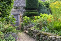 A gravel path next to a raised bed of dry stones. On top a planting of mixed flowering perennials. Beyond a wooden door in a wall near yew topiary at The Manor, Little Compton.