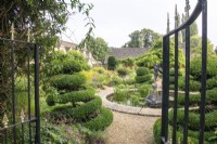 A view through gates into The Flower Garden with perennials, topiary and a circular pond at The Manor, Little Compton.