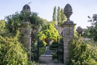 Wrought iron gates and stone pillars with decorative finials frame a view across The Flower Garden towards a summer house at The Manor, Little Compton.