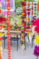 Marigold Garlands made with flowers, chilli peppers, fruits, pom-poms and bracelets in dinning area. The  RHS and Eastern Eye unity garden. Designer: Manoj Malde