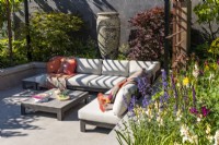 A sunken seating area with a comfortable corner sofa and low table surrounded by a perennial bed and two maple trees. A large ceramic vase decorates the background. Designer: Kevin Dennis