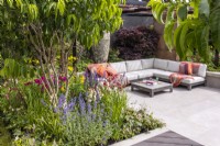 A view through Heptacodium leaves to a contemporary garden with sunken seating area with a modern corner sofa and coffee table. Beds planted with Acer palmatum and coloured perennials. Garden: Looms  and  Blooms, Designer: Kevin Dennis, Bord Bia Bloom 2023