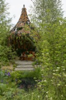 RHS Chelsea Flower Show 2023 - View over small pond, surrounded by Viburnum opulus and Iris sibirica, towards a steps up to a sitting area covered with ornate metal gazebo in The Boodles British Craft Garden Designed by Thomas Hoblyn