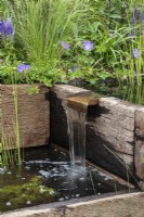 Reclaimed wood sleepers made into a water feature. Planting includes Geranium 'Johnston Blue' - Caroline and Peter Clayton - Get Started Gardens - Nurturing Nature in the City, RHS Hampton Court Palace Garden Festival.