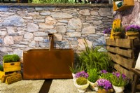 Rusted corten steel water tank by the wall stone, surrounded by herbs in pots on gravel surface and pallet in contemporary Italian courtyard. A water canal running on the gravel surface. 