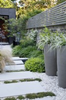 Large sandstone slabs lead to the zinc clad building; these are bordered on the right by Astelia chathamica in a tall pot, Pittosporum tobira 'Nanum' and clumps of Hakonechloa macra with Thymus praecox 'Albiflorus' running between the paving stones. 