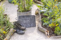 Edging made from broken tree branches. Old shoes next to the plaque and pot filled with peas. RHS Iconic Horticultural Hero Garden, Designer: Carol Klein