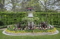 Urn with decorative layers of red, black and green foliage above lollipop trees underplanted with pink and white tulips and cordyline in a cylindrical flowerbed with concrete edging, Avenue Gardens, The Regent's Park, London, UK 