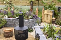 View picnic area on wooden deck with modern chiminea and mini pond to a raised bed made of  Connemara walling system in a woodland inspired garden. Planted with  Leucanthemum superbum, Salvia Nemorosa and aquatic plants. June
