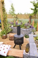 View picnic area on wooden deck with modern chiminea to a raised bed made of  Connemara walling system. Planted with perennials in a woodland inspired garden surrounded by a wooden planks fence. June
Designer: Mary Anne Farenden. Bord Bia Bloom, Super Garden, Dublin, Ireland.