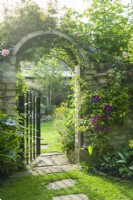 Wrought iron garden gate in a brick wall clothed with roses and clematis. Stepping stones leading across a neatly mown lawn. June