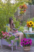 A young woman enjoys reading a magazine on a decked summer terrace with containers planted with pelargoniums and Impatiens.