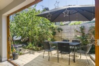 A small sun filled patio garden designed as an outdoor living space with large parasol, garden table and chairs and a pair of hanging seats. June.