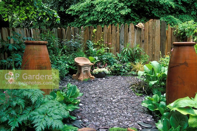 Small patio garden with slate chippings, foliage planting, rustic wooden fence, and terracotta urns and seat