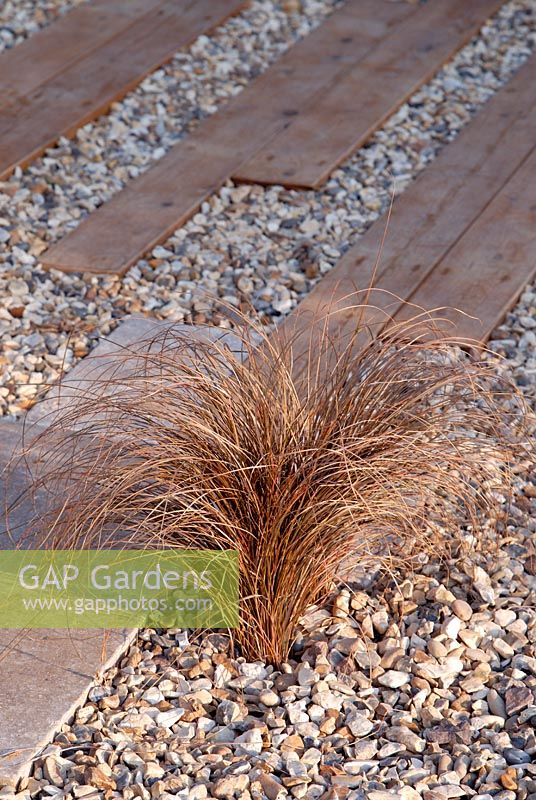 Carex flagellifera planted in gravel with wooden and stone paving in 'The Homebase Living Room' garden 
