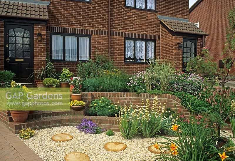 Front garden of modern house with retainig wall, steps and raised borders of Geranium, Persicaria affinis and Grasses. Sisyrinchium striatum, Anthemis and Campanula growing in gravel. Small sorbus tree underplanted with Hemerocallis. Circular paving slabs set in gravel. Containers of evergreens