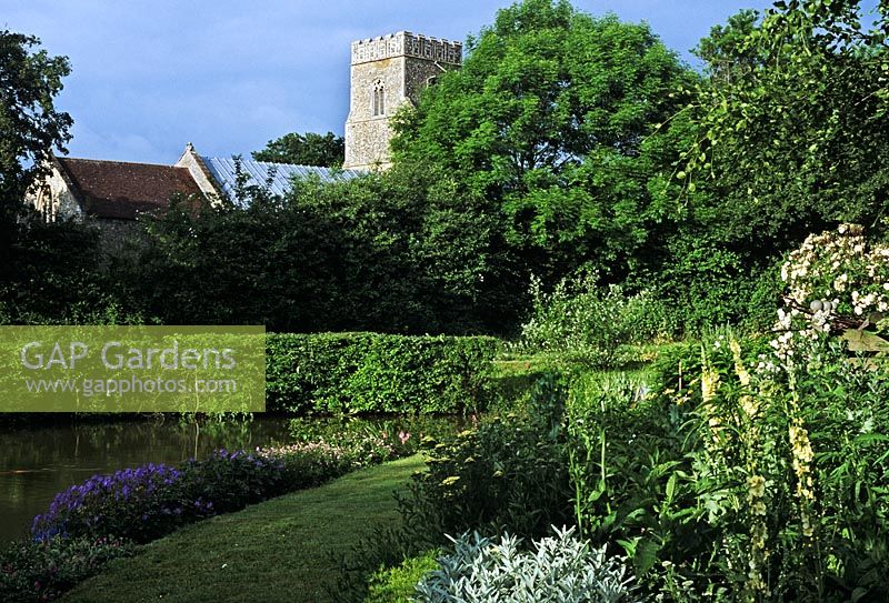 Country garden with grass path leading to moat. Village church in background - Bedfield Hall, Suffolk