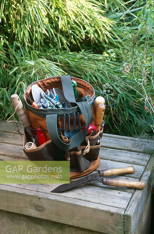 View of canvas garden tool bag with handle -Pockets holding tools including secateurs, pruning knife, pruning saw, labels, trowel, fork, ball of string and gloves - Small garden shears lying beside it on wooden bench
