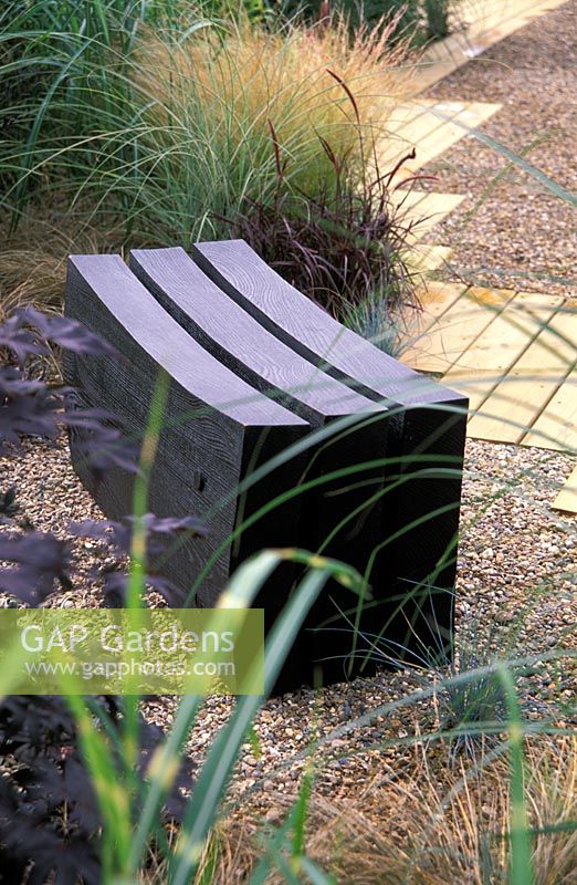 A black, curved, timber seat set within gravel with a timber path and planting of grasses