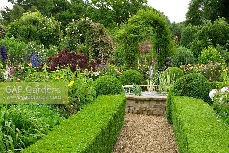 Box edged beds containing profuse planting in June with formal raised pool and arches of climbers 