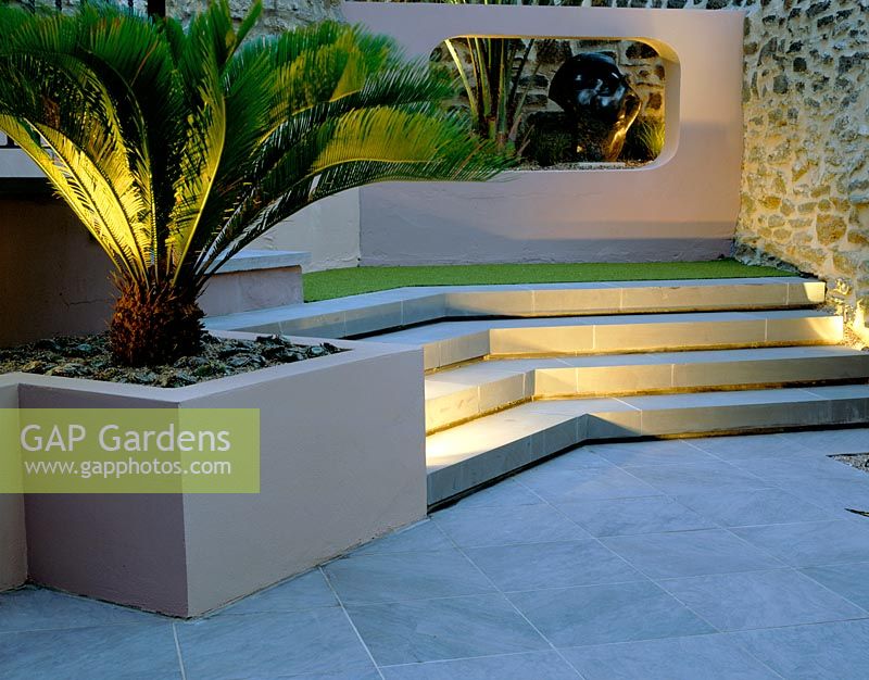 Raised bed with Cycas revoluta, concrete aperture with Butia capitata and sculpture lit up at night in small courtyard