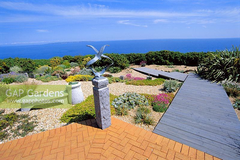 Seaside garden in guernsey with view out to sea, brick patio, gravel, wooden boardwalk, chinese granite seat and 'Seagulls' sculpture by Guy Portelli