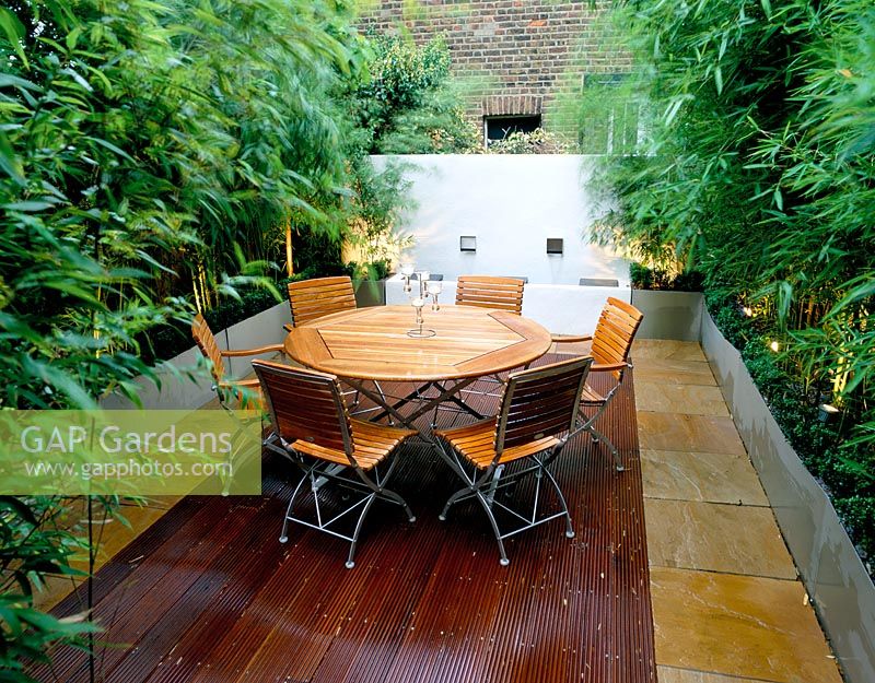 Roof terrace with decking at night - Wooden table and chairs, galvanised metal containers with bamboo and whitewashed wall 