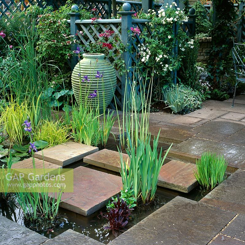Square stepping stones across sunken pond with large verdigris stoneware pot behind