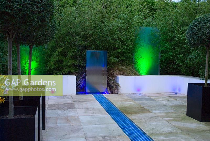 Small contemporary paved urban garden with night lighting on stainless steel water feature and metal grille rill illuminated