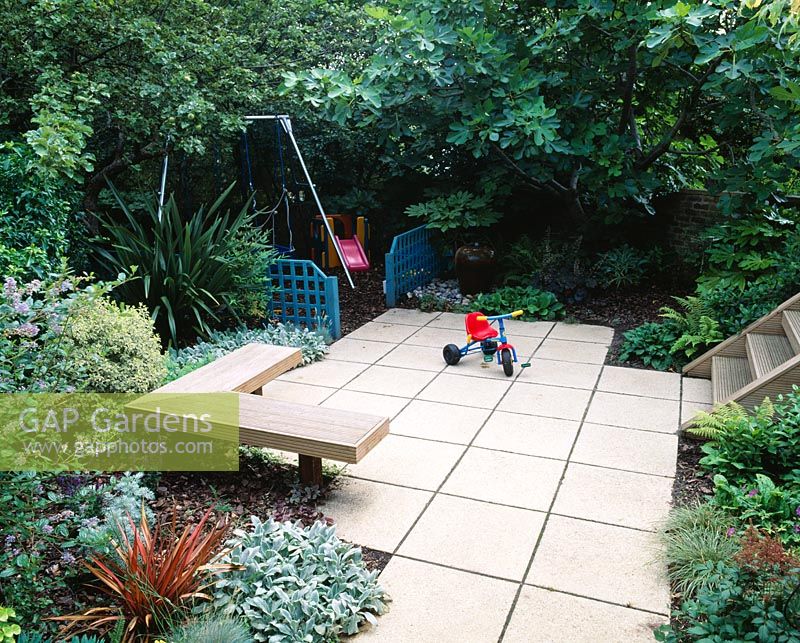Childrens garden with swings with bark beneath, blue trellis screens, paving, wooden seat, Phormium and Ficus - fig tree