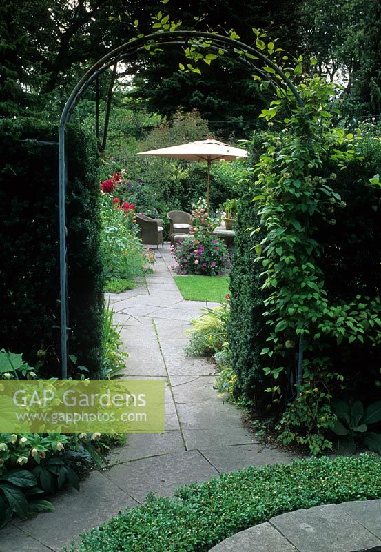 Arch in hedge above paving slab path with view to table chairs and umbrella beyond. 


