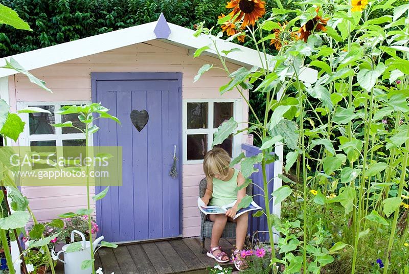 Young girl sitting reading on porch of painted wendy house with border of sunflowers