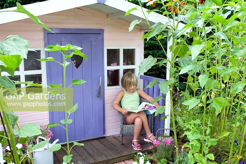 Young girl sitting reading on porch of painted wendy house with border of sunflowers 