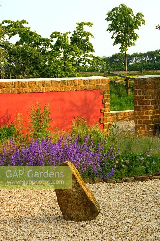 View across gravel garden to rock and red wall, border with Nepeta 'Walkers Low'