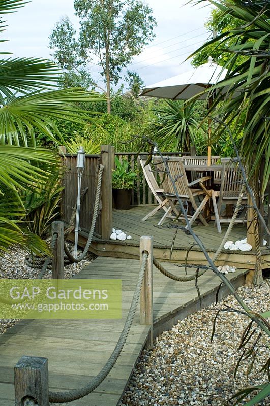 Garden design inspired by Daintree forest, Australia. Boardwalk with rope edging leading to raised deck with table and chairs. Planting of Chusan palm, Phormium and Eucalyptus - Felsted, Essex