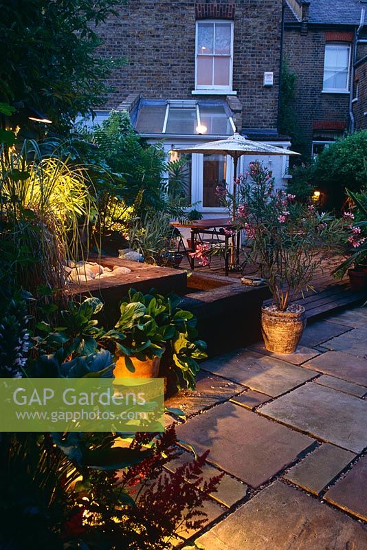 Small town back garden at night with paved patio area with table and chairs.  
Railway sleeper raised water feature and container of Oleander - London 