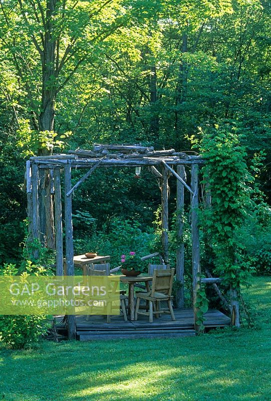 Table and chairs on shaded deck patio  beneath wooden arbour made with rustic poles - NY State, USA 