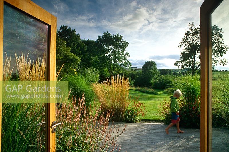 Double glass doors looking out to garden with lawn and limited palette perennials planting with grasses. Boy walking on wooden deck terrace - Fawler Copse