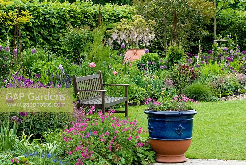 Pink, purple and blue border with Salvia, Allium, Geranium, Erysimum, Iris, Papaver and Wisteria with containers, bench and lawn.